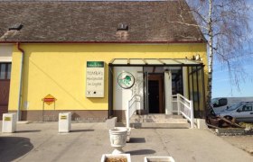 Gelbes Gebäude mit Schild 'Tomba's Heurigenschenke'. Eingang mit Treppe und Glasüberdachung.