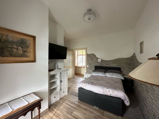 Ein gemütliches Zimmer mit einem Einzelbett, einem Fernseher an der Wand und einem kleinen Tisch. Helle Wände und Holzboden schaffen eine einladende Atmosphäre.