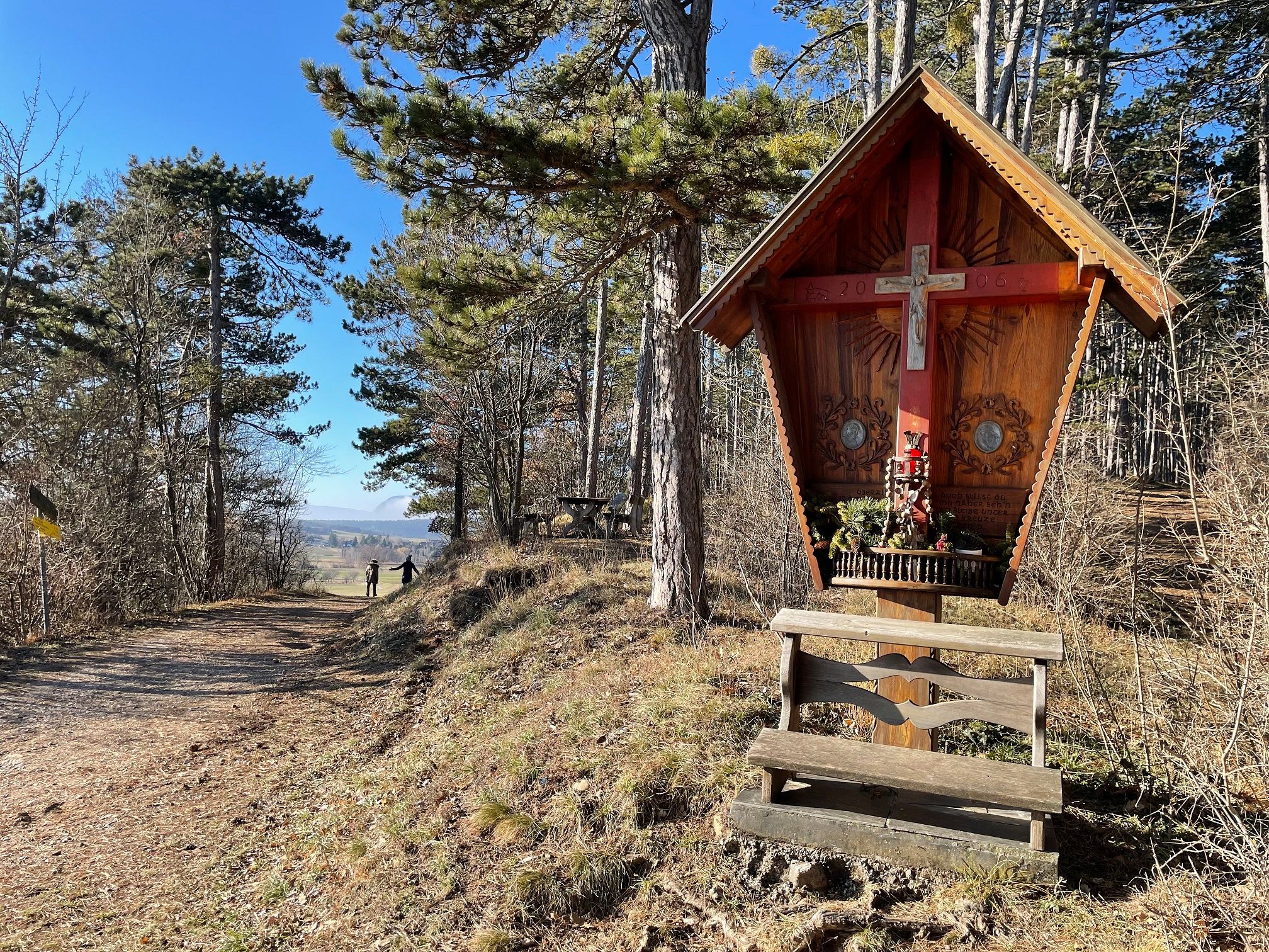 Holzkreuz mit Bank in einem Waldgebiet, sonniger Tag.
