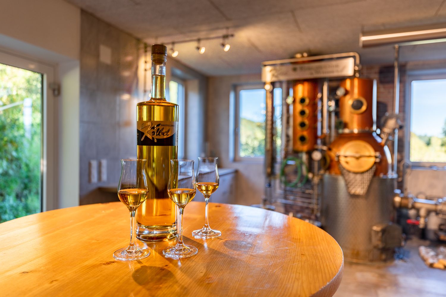 Eine Flasche und drei halb gefüllte Gläser stehen auf einem runden Holztisch vor einer Brennanlage.