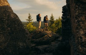 Zwei Wanderer stehen zwischen imposanten Felsformationen und genie&szlig;en den weiten Blick auf die Waldlandschaft im goldenen Morgenlicht im Waldviertel.