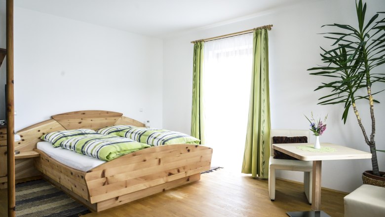 Helles Zimmer mit Holzbett, gr&uuml;n-wei&szlig; gestreifter Bettw&auml;sche, Tisch mit Vase und Pflanze, gr&uuml;ne Vorh&auml;nge.