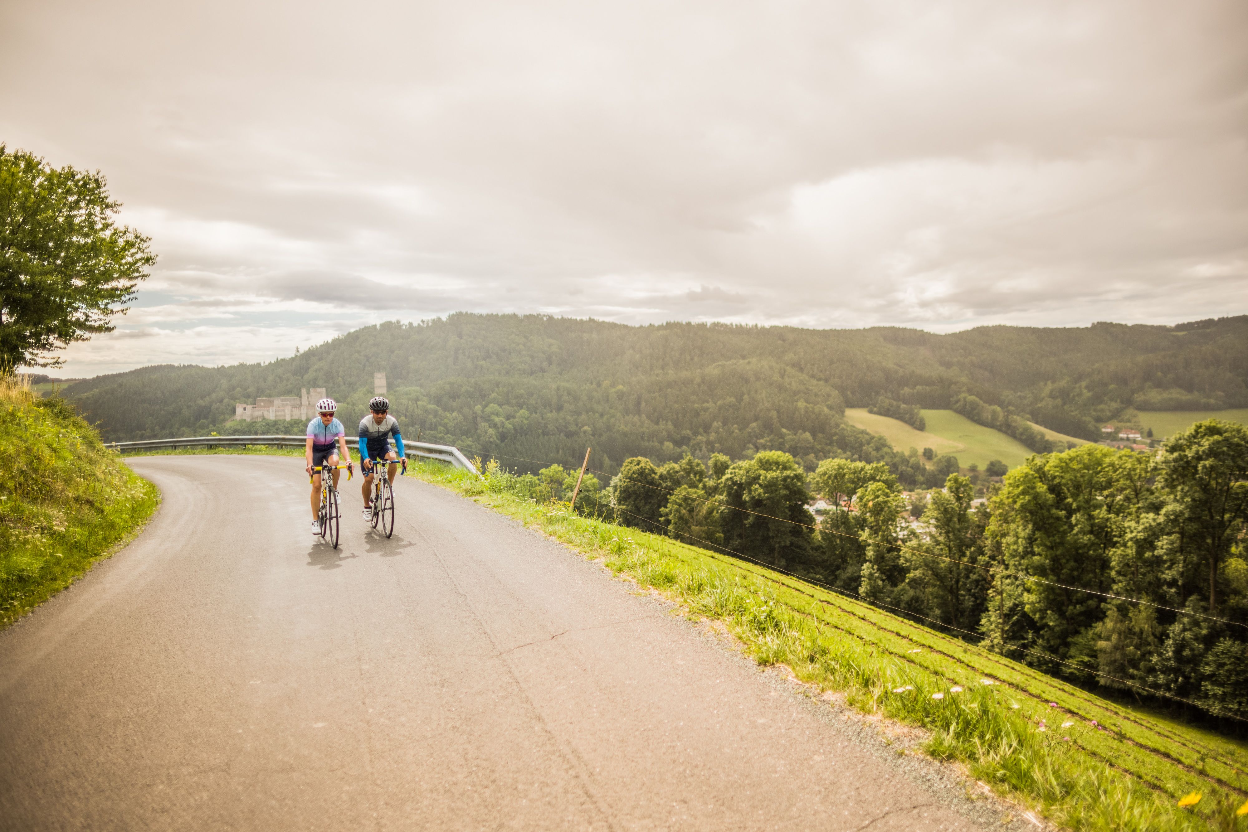 Zwei Personen am Fahrrad auf einer Straße in hügeliger Landschaft mit einer Burg im Hintergrund.
