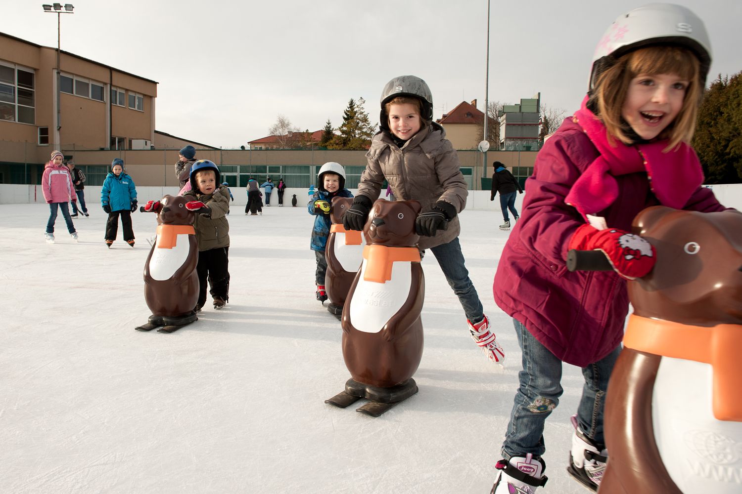 Kinder beim Schlittschuhlaufen mit Pinguin-Hilfen auf einer Eisbahn.