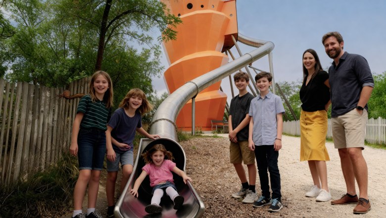 Kinder spielen auf einer Rutsche im Freien mit einem großen orangefarbenen Turm im Hintergrund.
