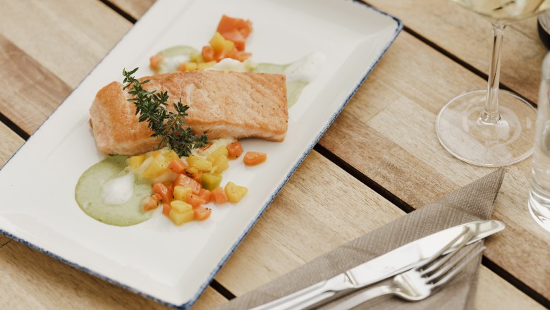 Ein Teller mit gebratenem Lachs auf Gem&uuml;se und gr&uuml;ner Sauce, dekoriert mit einem Kr&auml;uterzweig, auf einem Holztisch mit Besteck und Weinglas.