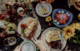Ein Tisch mit verschiedenen Gerichten, darunter Salat, Brot mit Belag und Wein, dekoriert mit Sonnenblumen.