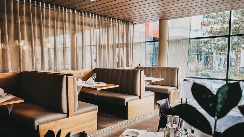 Innenansicht eines eleganten Restaurants mit Holzm&ouml;beln und gro&szlig;en Fenstern.