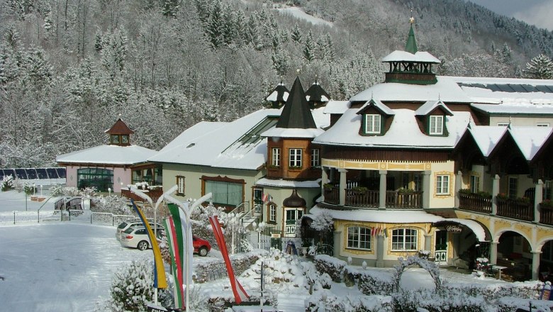Ein verschneites Hotel im alpinen Stil mit T&uuml;rmen und Balkonen, umgeben von schneebedeckten B&auml;umen und H&uuml;geln.