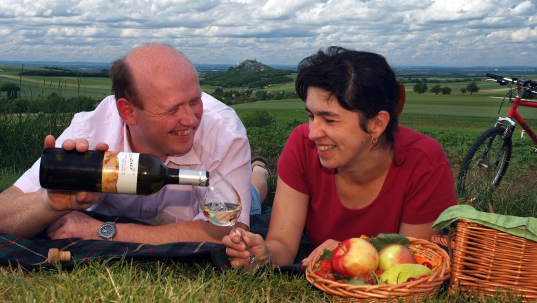 Ein Paar beim Picknick auf einer Wiese mit Wein und Obstkorb, im Hintergrund eine h&uuml;gelige Landschaft und ein Fahrrad.