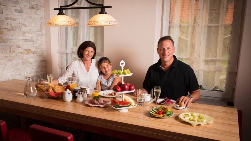 Familie beim Frühstück an einem Holztisch mit Obst und Getränken.