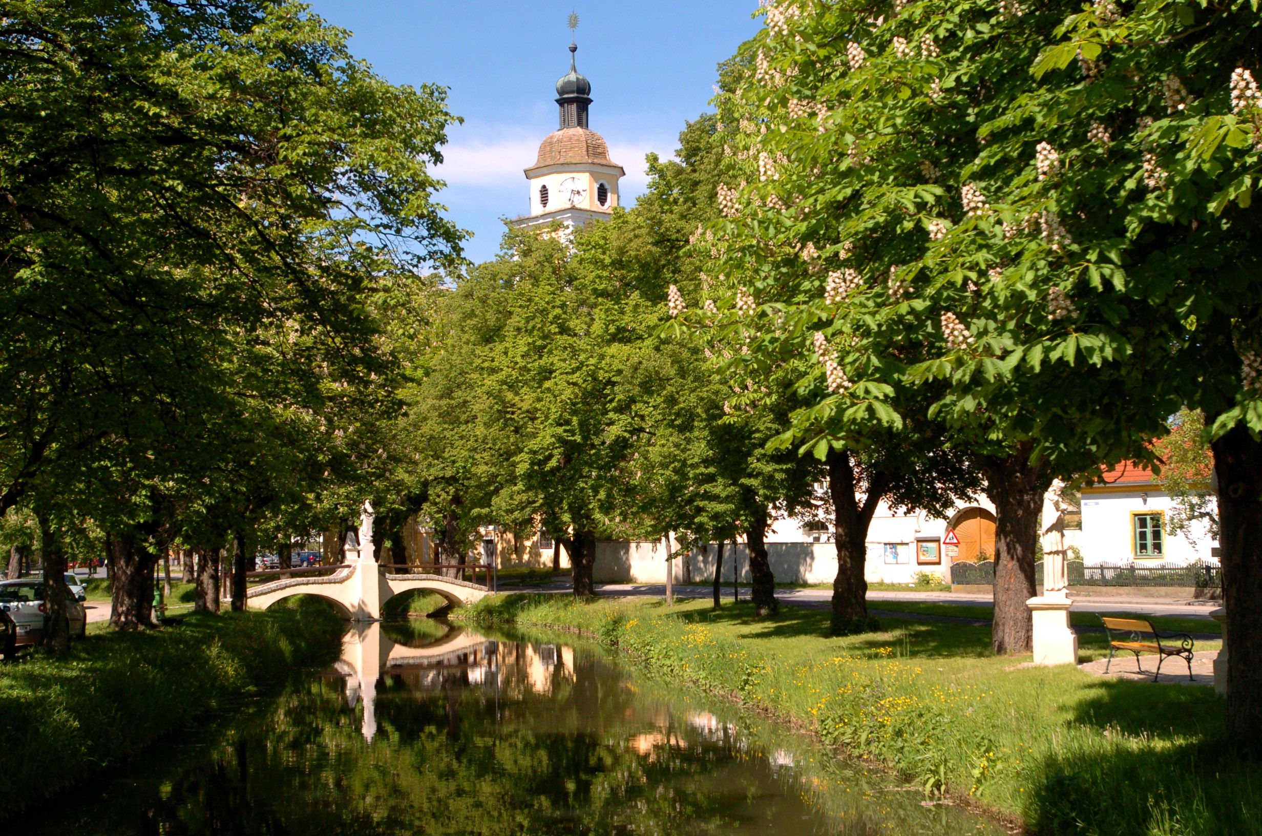 Ein idyllischer Park mit einem kleinen Fluss, einer Brücke und einem Kirchturm im Hintergrund.