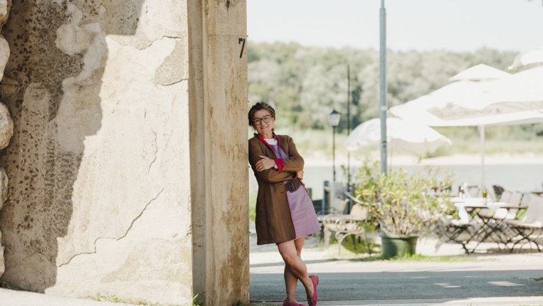 Wirtin Michaela Brenner-Melchior, © Niederösterreich Werbung/Michael Reidinger Frau lehnt lächelnd an einer Mauer, im Hintergrund Terrasse mit Tischen, Stühlen und Sonnenschirmen.