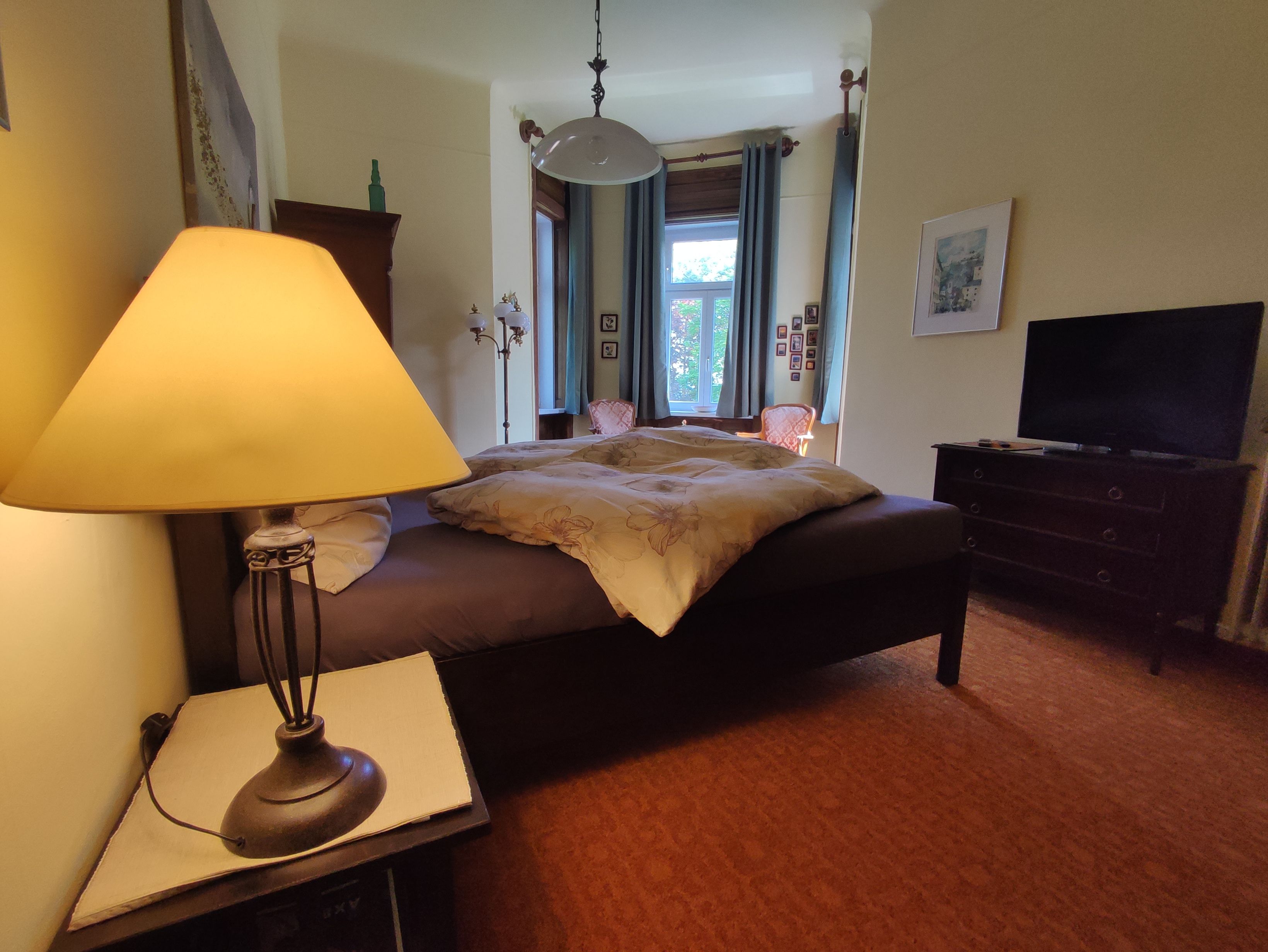 Gemütliches Schlafzimmer mit Doppelbett, Nachttischlampe, Fenster und Fernseher.