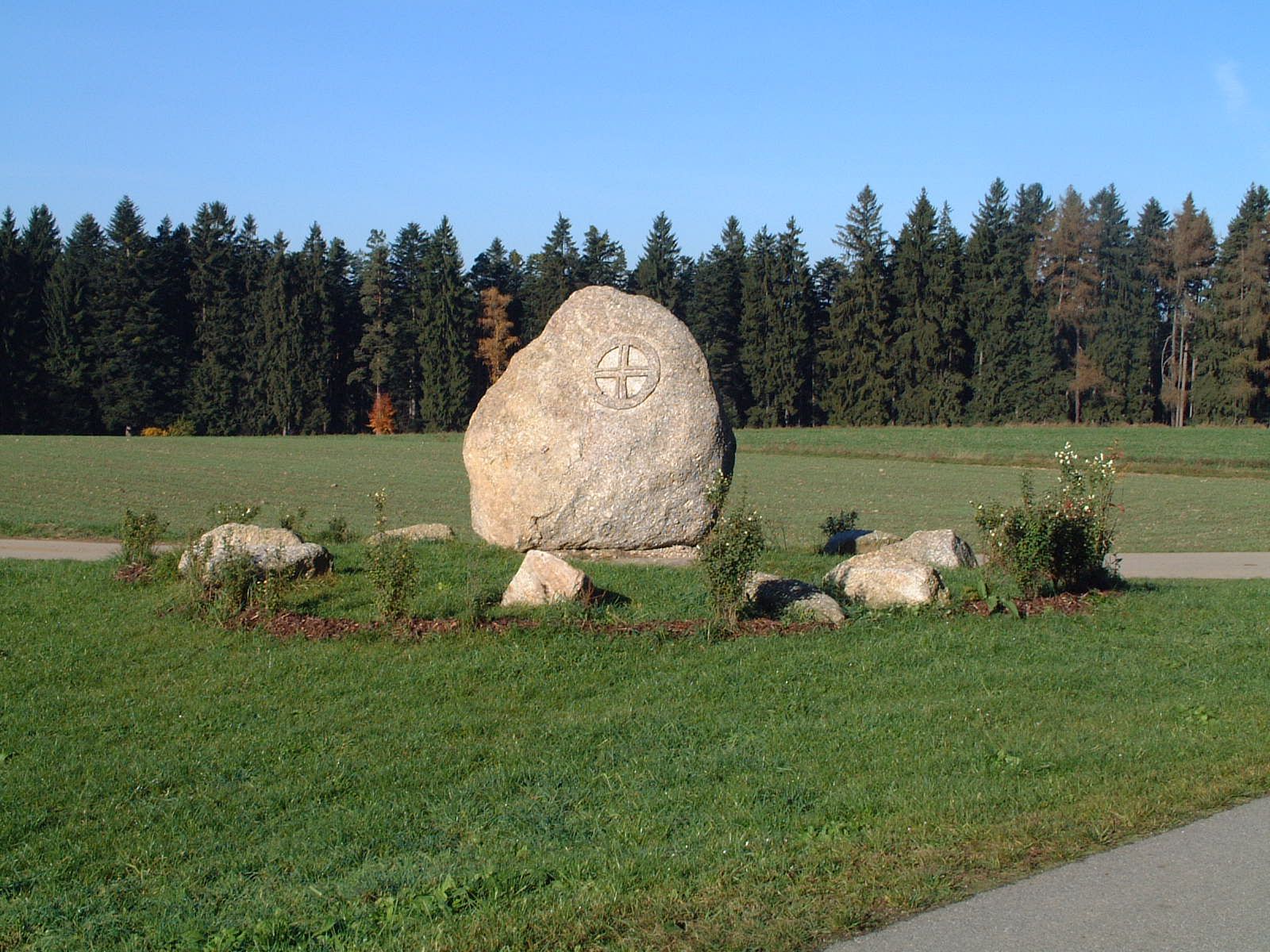 Großer Stein mit eingraviertem Symbol auf einer Wiese, umgeben von kleineren Steinen und Pflanzen, im Hintergrund ein Wald.