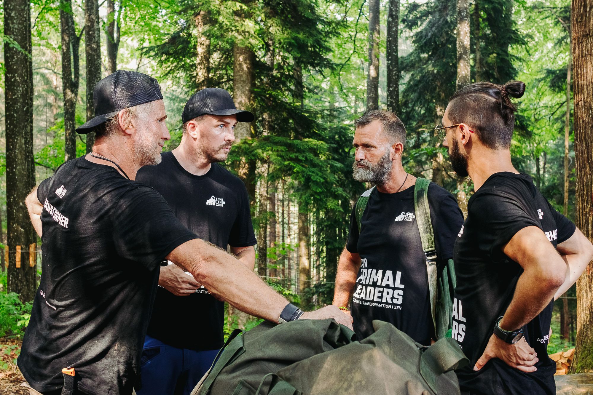 Vier Männer in schwarzen T-Shirts stehen im Wald und unterhalten sich.