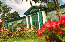 Ein gr&uuml;nes Geb&auml;ude mit gro&szlig;en Fenstern, umgeben von bunten Blumen und Palmen, unter einem blauen Himmel.