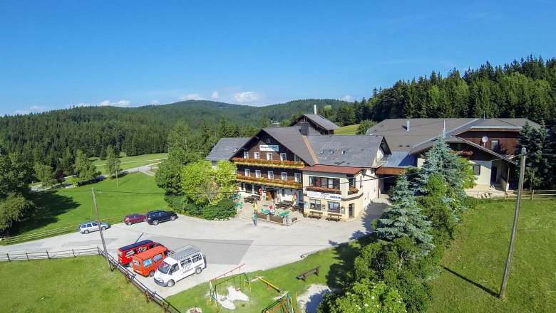 Alpengasthof Postl in einer gr&uuml;nen, bewaldeten Landschaft mit mehreren geparkten Autos und einem Spielplatz im Vordergrund.