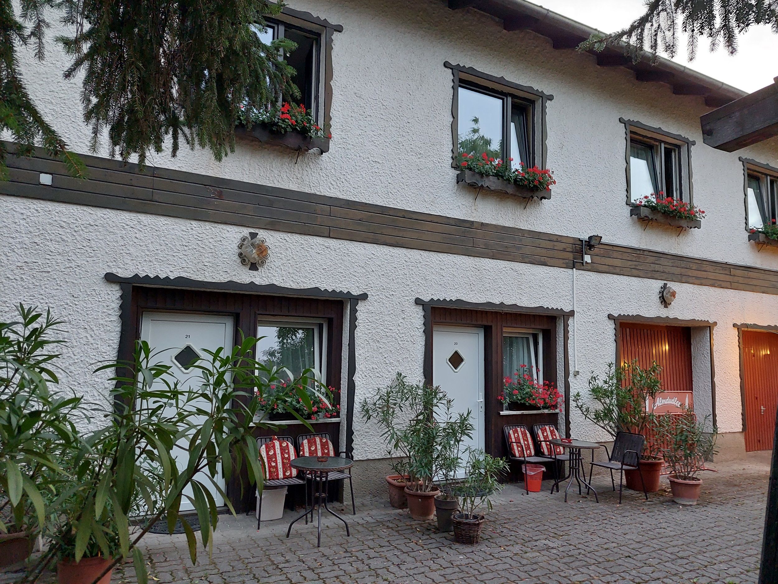 Außenansicht eines zweistöckigen Gebäudes mit weißen Wänden, Blumenkästen und Sitzgelegenheiten vor den Türen.