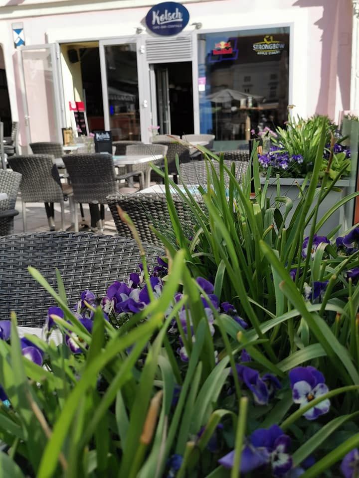 Ein Schanigarten mit Korbstühlen und Tischen vor einem Café, umgeben von blühenden Blumen und Pflanzen.