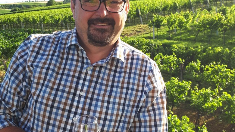 Ein Mann im karierten Hemd steht mit einem Weinglas in der Hand vor einem Weinberg.