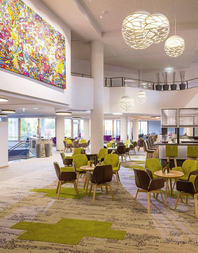 Moderne Hotellobby mit Teppichboden, runden Tischen, grünen Stühlen, großen Lampen und einem Wandbild.