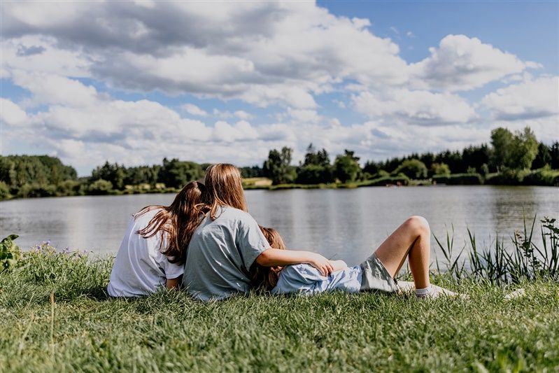 Drei Personen sitzen auf einer Wiese am See und schauen aufs Wasser.