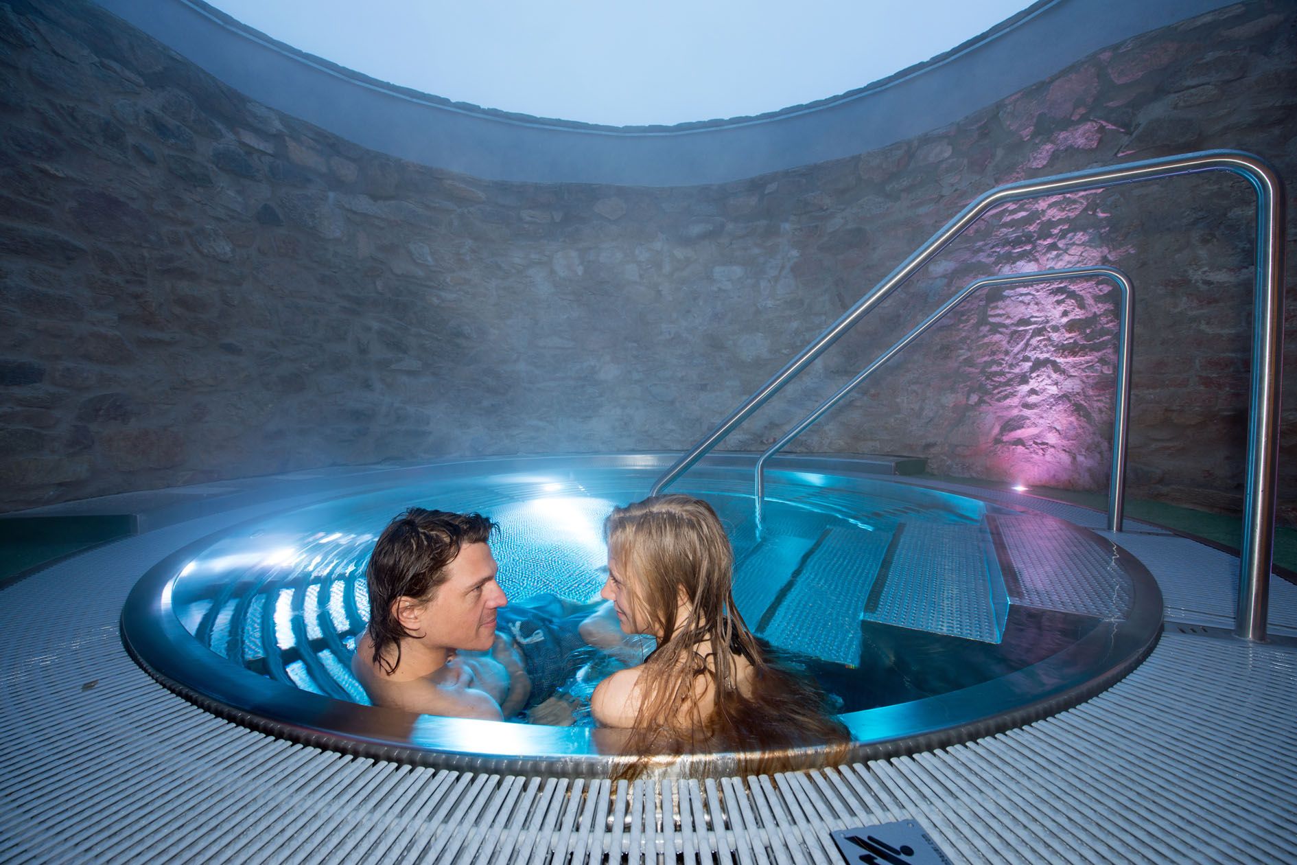 Ein Paar entspannt in einem runden Whirlpool mit blauer Beleuchtung und Steinwänden.