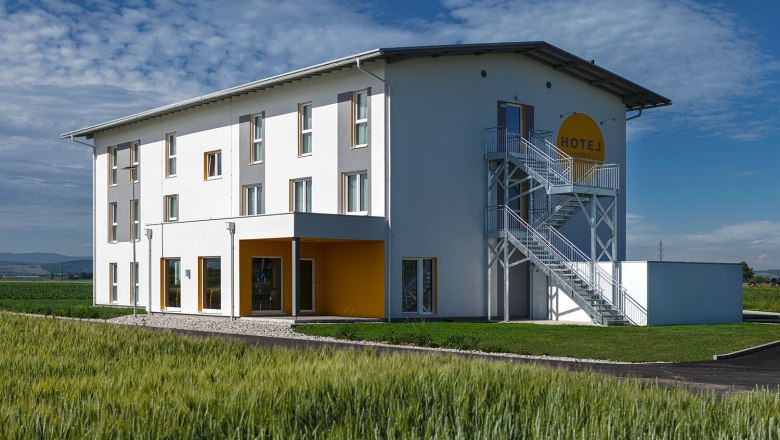 Aussenansicht Hotel Tullnerfeld, © Hotel Tullnerfeld Außenansicht eines modernen Hotels mit weißer Fassade und gelben Akzenten, umgeben von Feldern.
