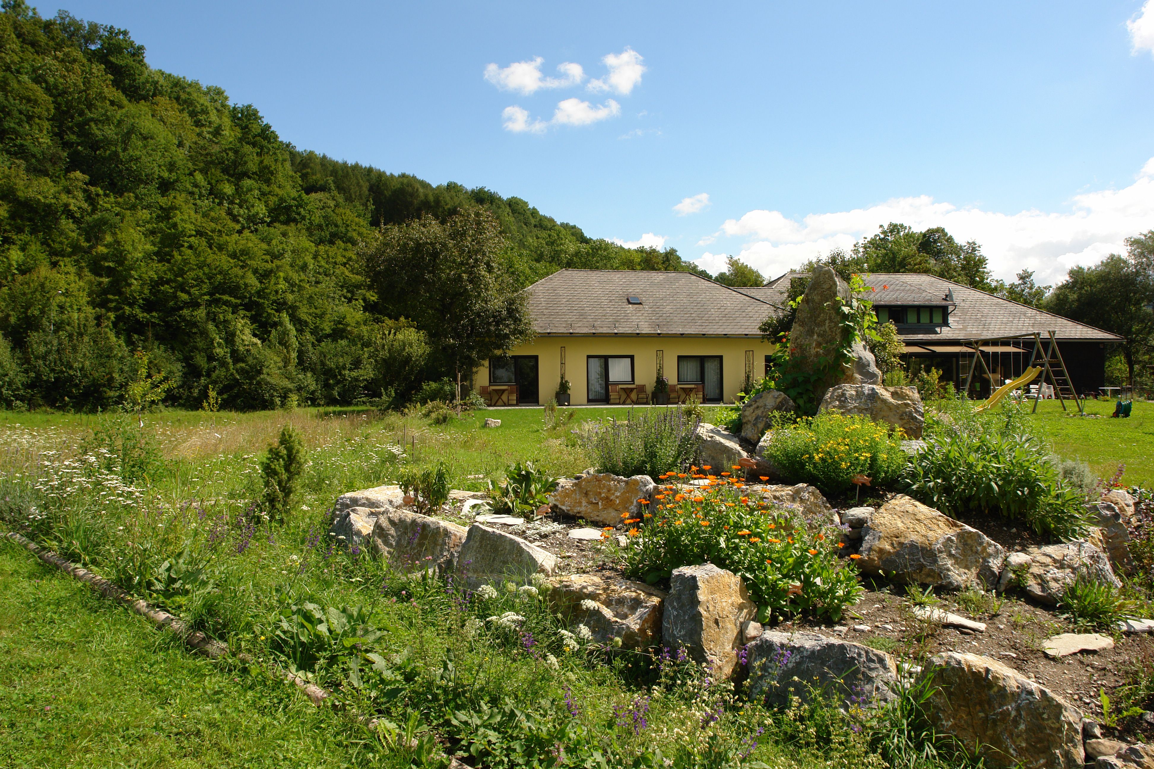 Ein gelbes Haus mit Garten und Felsen im Vordergrund, umgeben von Bäumen.