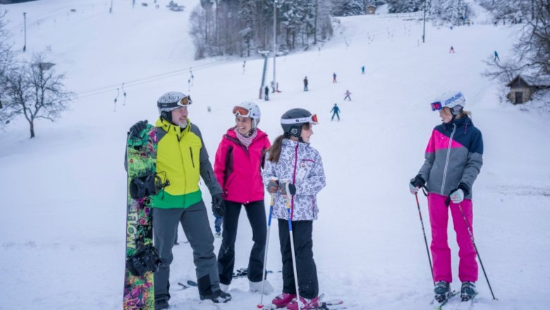 Gruppe von Skifahrern und einem Snowboarder auf einer verschneiten Piste mit Skilift im Hintergrund.