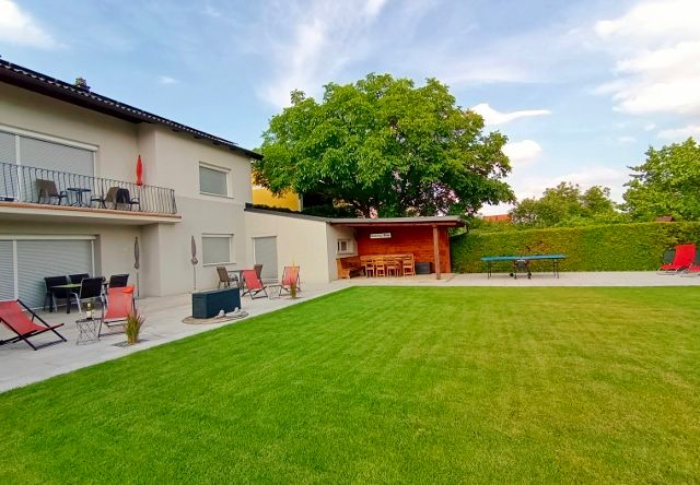 Ein gepflegter Garten mit Rasen, Terrasse, Liegestühlen und einem großen Baum neben einem modernen Gebäude.