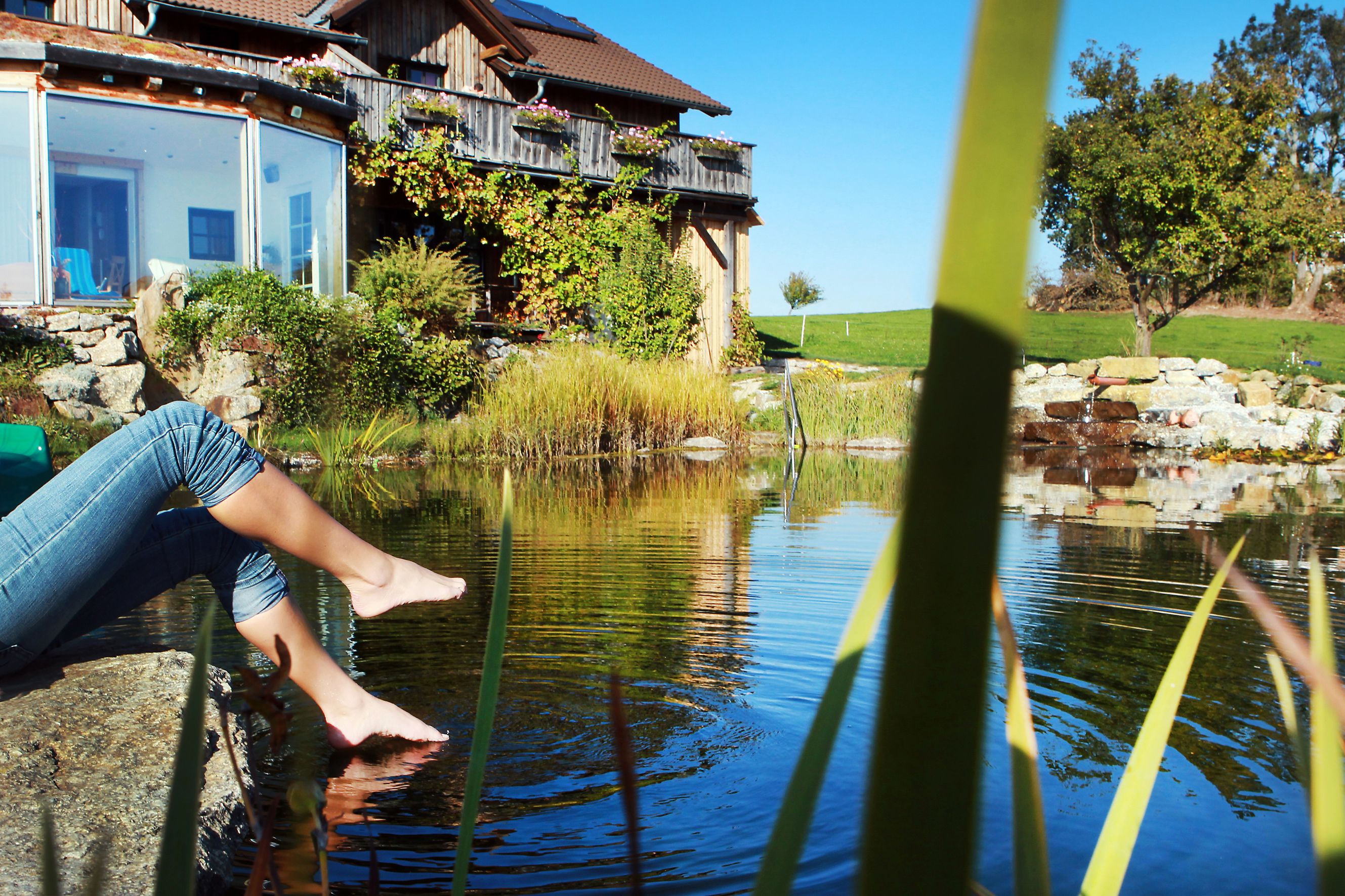 Person sitzt am Teichrand mit den Füßen im Wasser, vor einem Bauernhaus mit Garten.