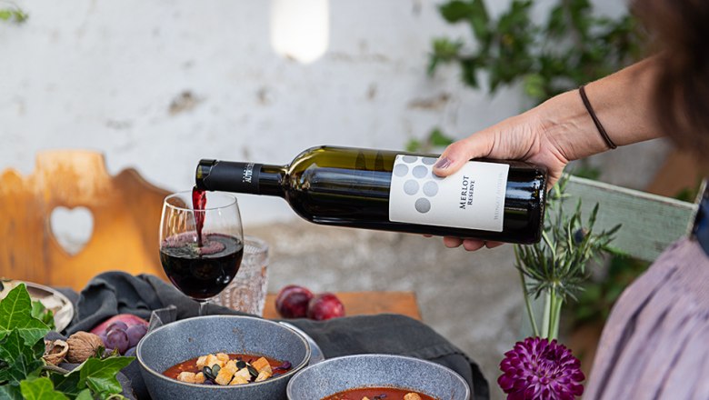 Eine Hand schenkt Rotwein in ein Glas neben zwei Schalen Suppe auf einem Tisch.