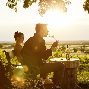 Mann trinkt von einem Weinglas und sitzt auf einer Holzbank mit einer Frau im Weingarten.