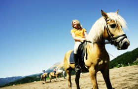 Eine Frau reitet auf einem braunen Pferd mit blonder Mähne auf einem Reitplatz, umgeben von Bergen und blauem Himmel.