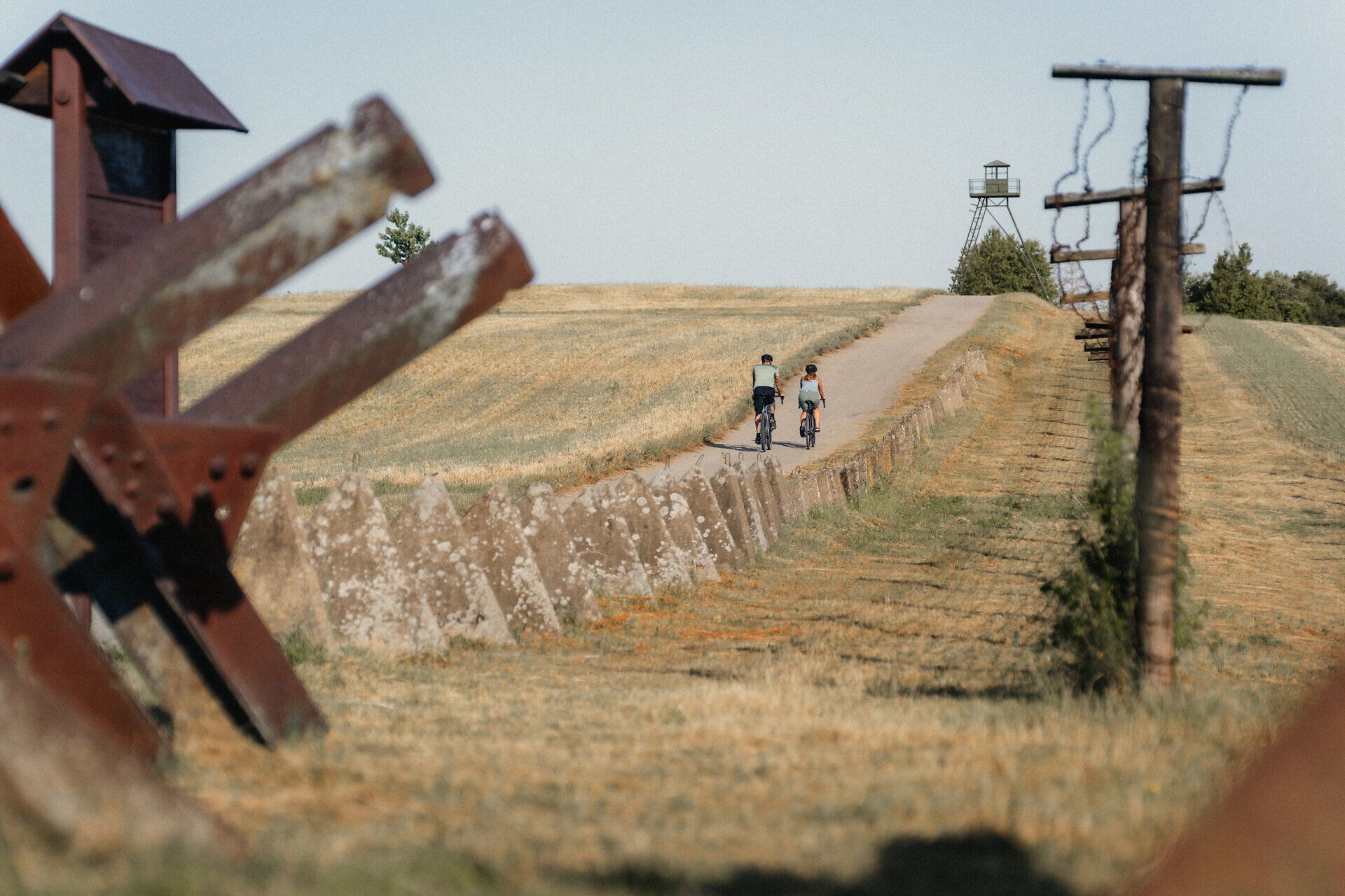 Zwei Radfahrer auf einem Weg neben einem alten Grenzzaun und einem Wachturm.