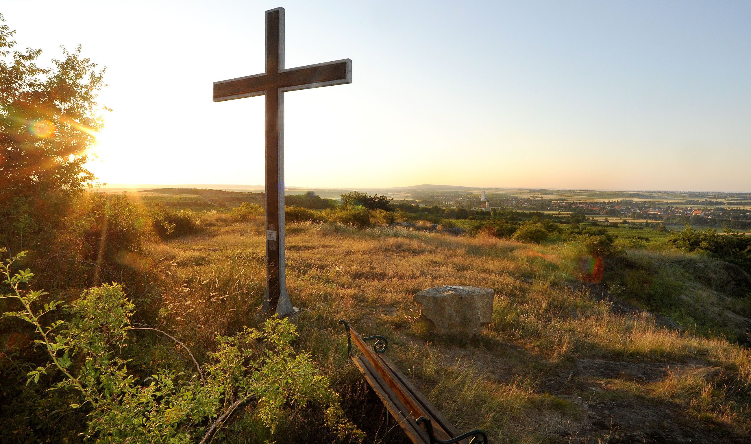 Ein Kreuz auf einem Hügel bei Sonnenuntergang mit Blick auf eine weite Landschaft und ein Dorf im Hintergrund.