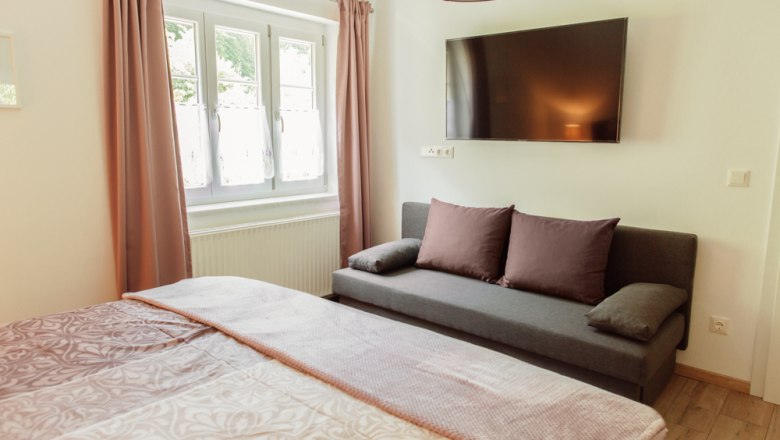 Innenansicht eines Schlafzimmers mit Bett, Sofa, Fenster und Wandfernseher.