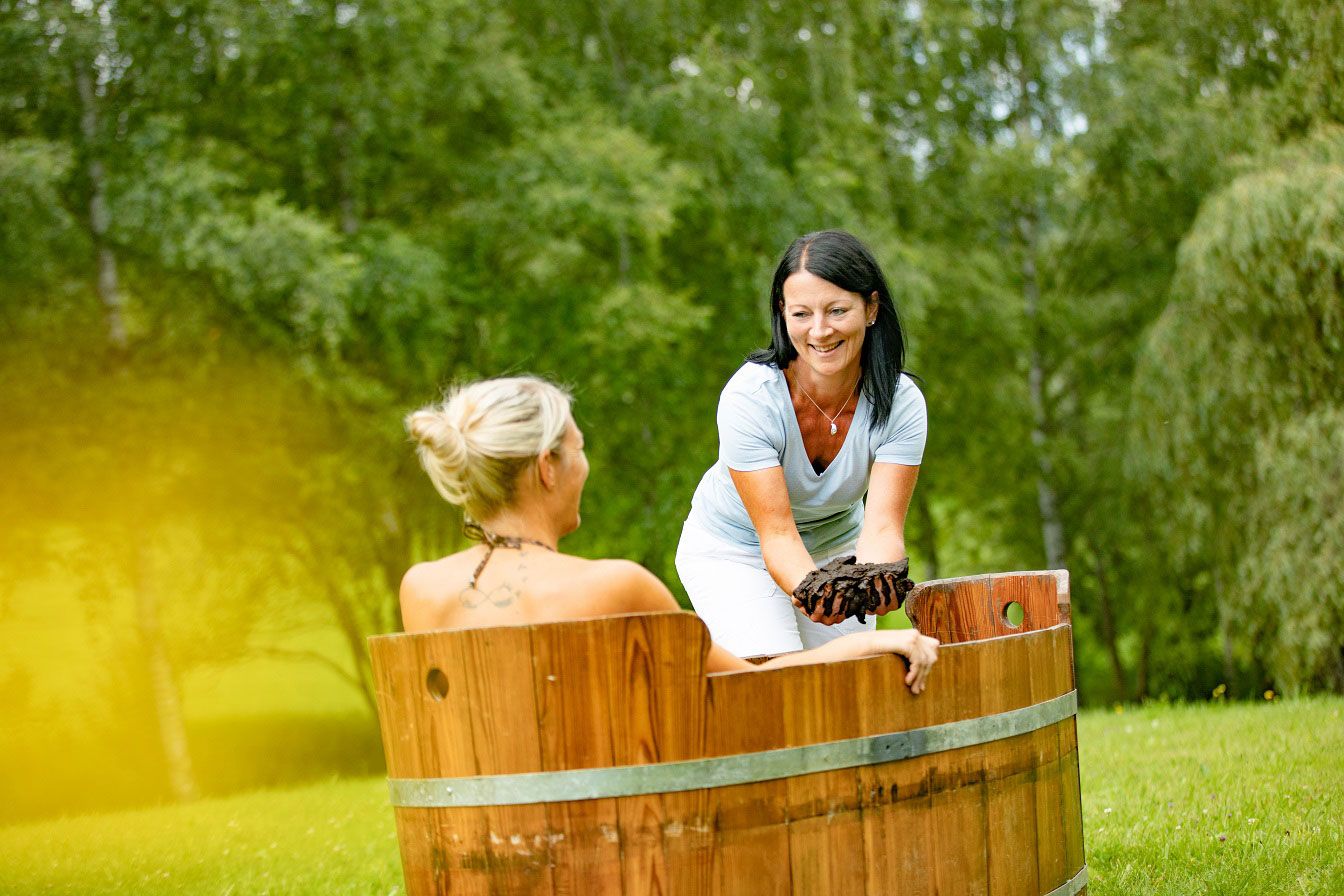 Zwei Frauen bei einer Moorbehandlung im Freien, eine sitzt in einer Holzwanne, die andere reicht ihr Moor.