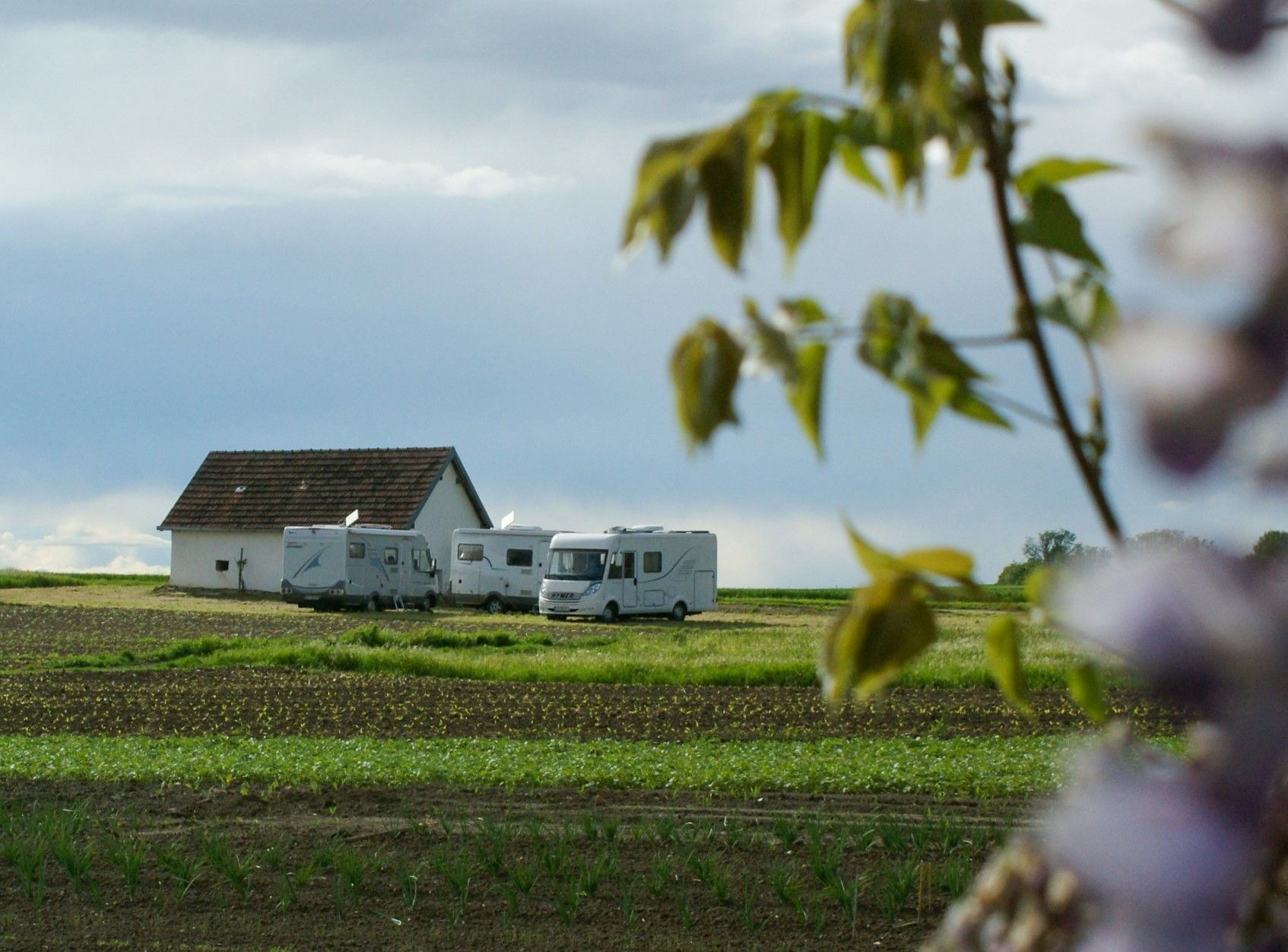 Wohnmobile auf einem Feld neben einem kleinen Gebäude, umgeben von grüner Landschaft und einem bewölkten Himmel.