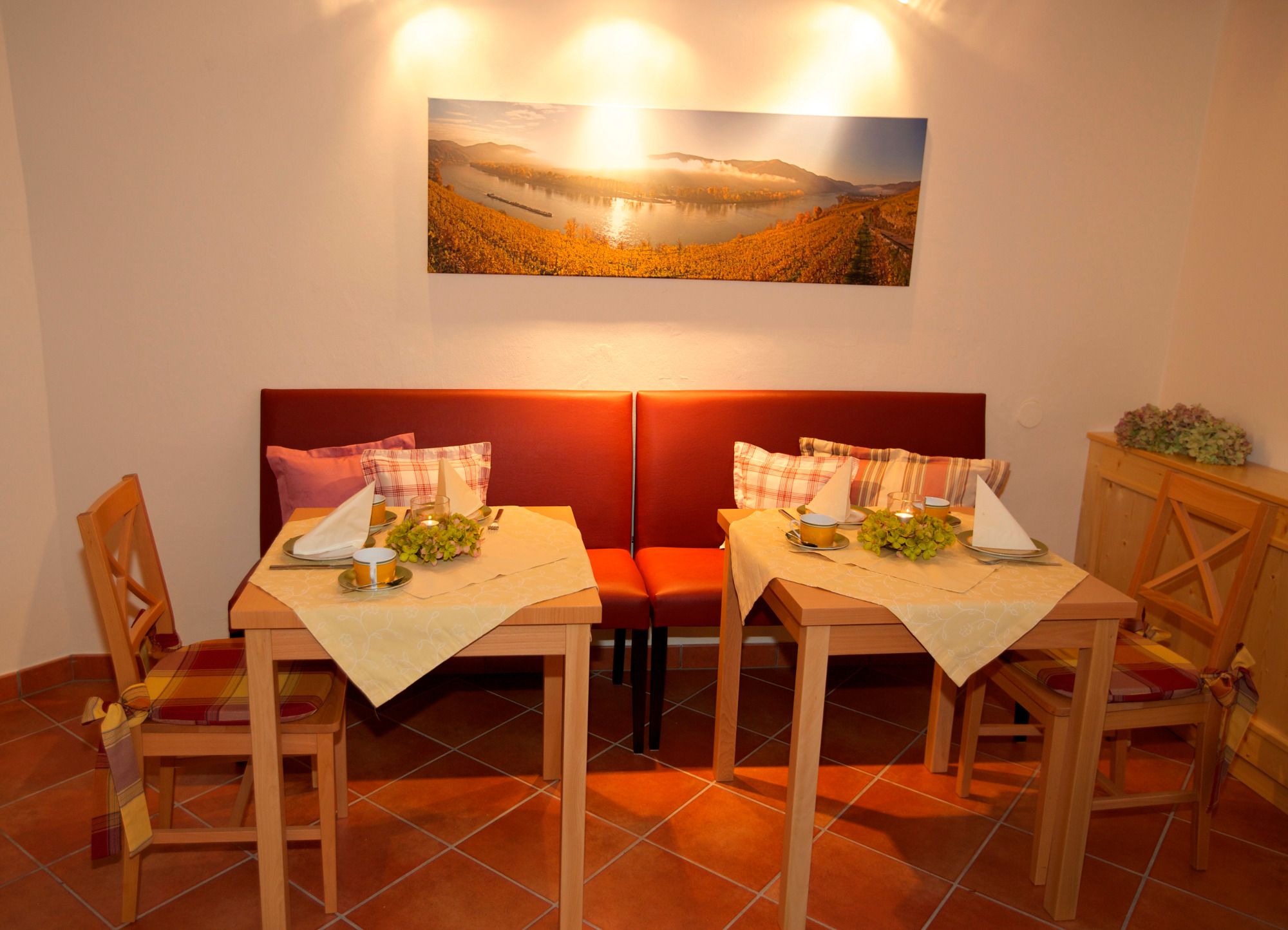 Gemütlicher Frühstücksraum mit zwei gedeckten Tischen, roten Sitzbänken und einem Landschaftsbild an der Wand.