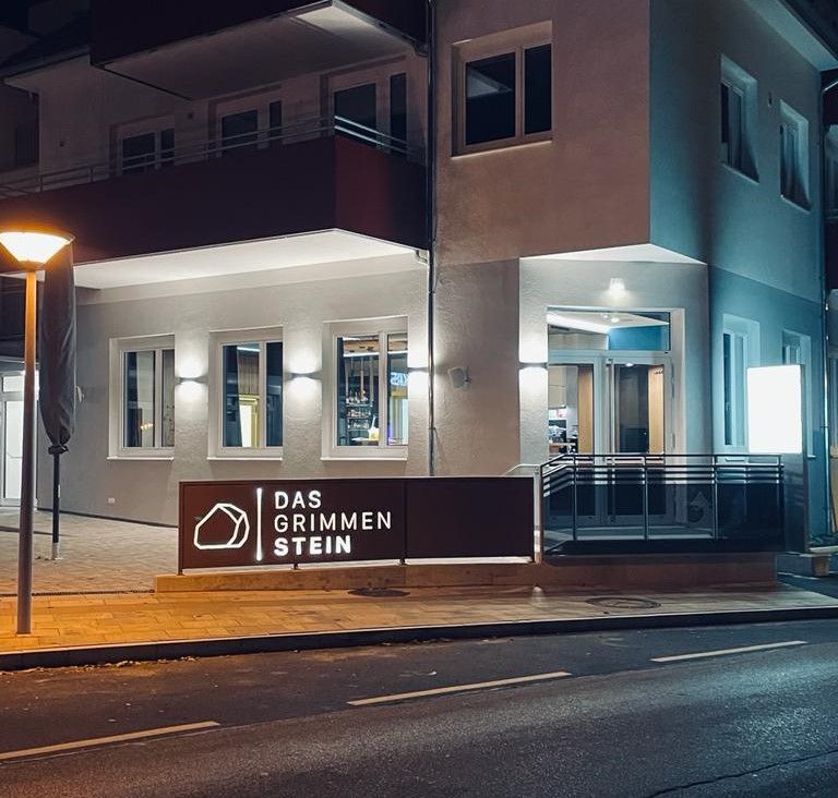 Außenansicht eines beleuchteten Gebäudes bei Nacht mit dem Schild 'Das GrimmenStein'.