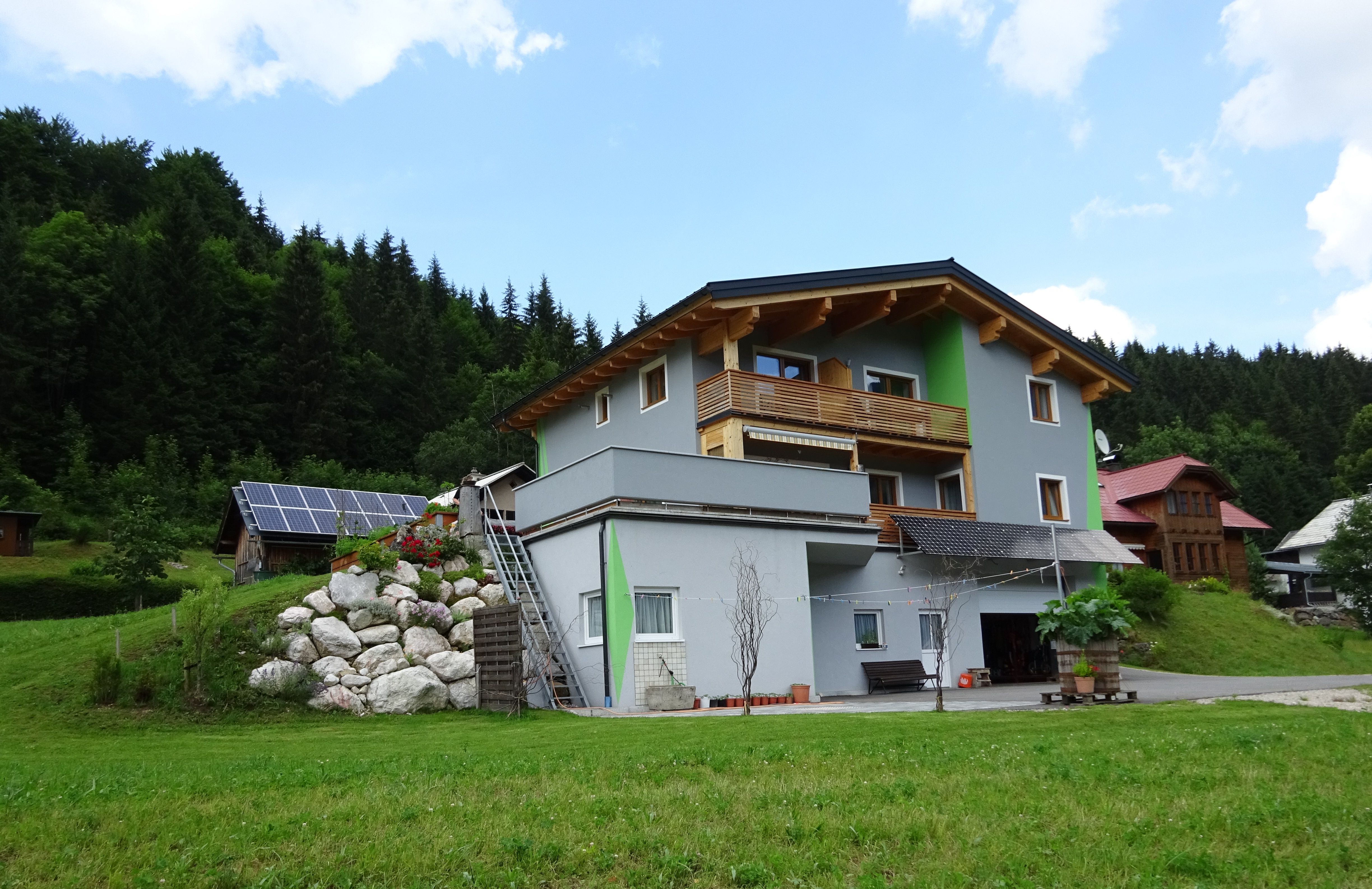Ein modernes Haus mit Holzverkleidung und Solarpanelen in einer grünen, bewaldeten Umgebung.