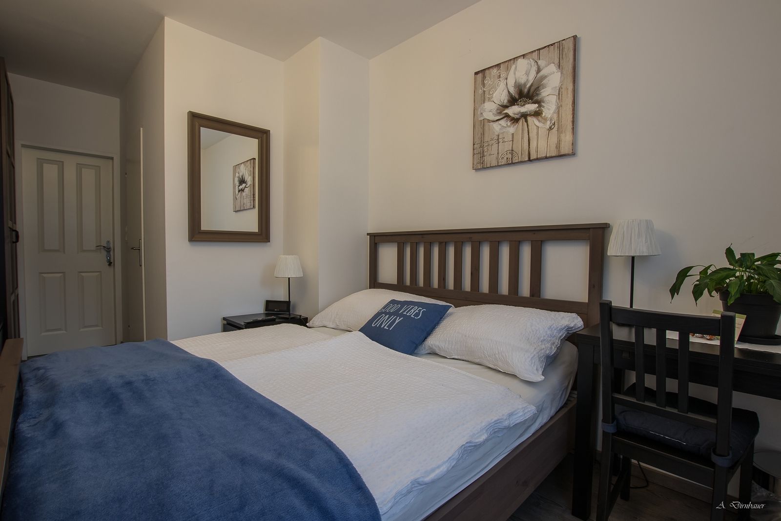 Gemütliches Doppelzimmer mit Holzbett, blauer Decke und Dekokissen. Ein Spiegel und ein Blumenbild an der Wand, daneben ein Schreibtisch mit Pflanze.