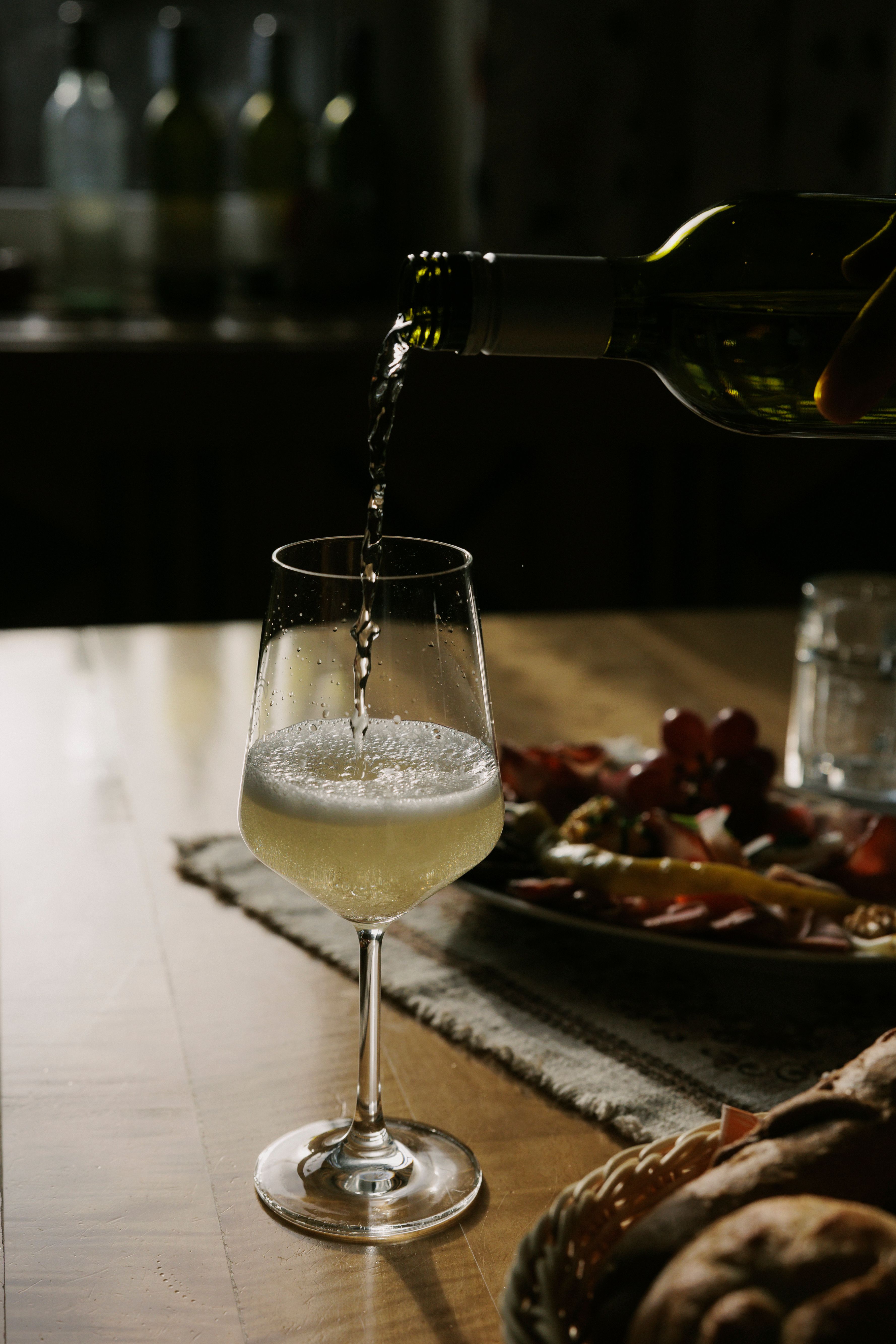 Ein Glas Weißwein wird auf einem Tisch eingeschenkt, im Hintergrund ein Teller mit Snacks.