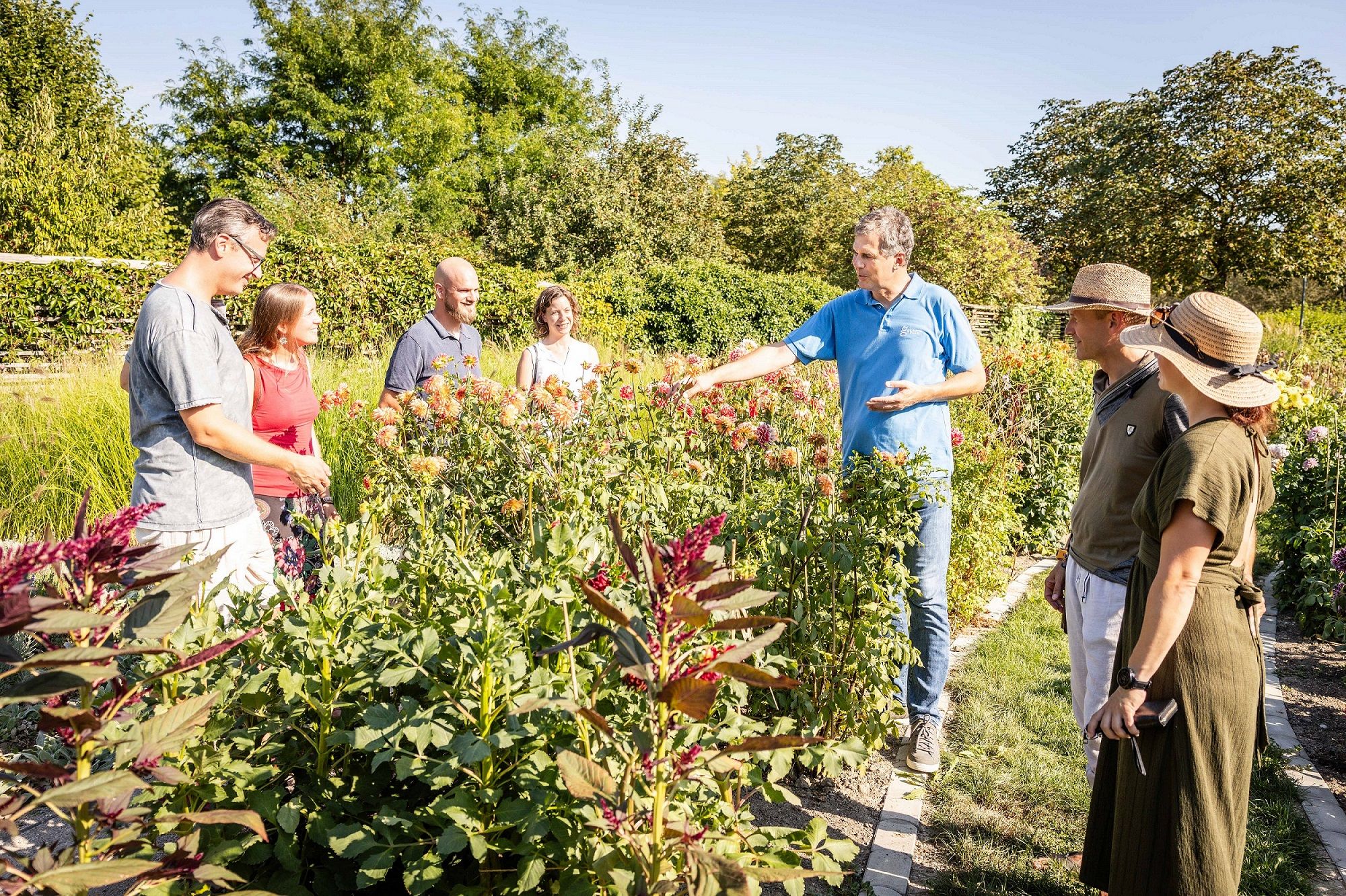 Eine Gruppe von Menschen steht in einem blühenden Garten und hört einem Mann zu, der etwas erklärt.