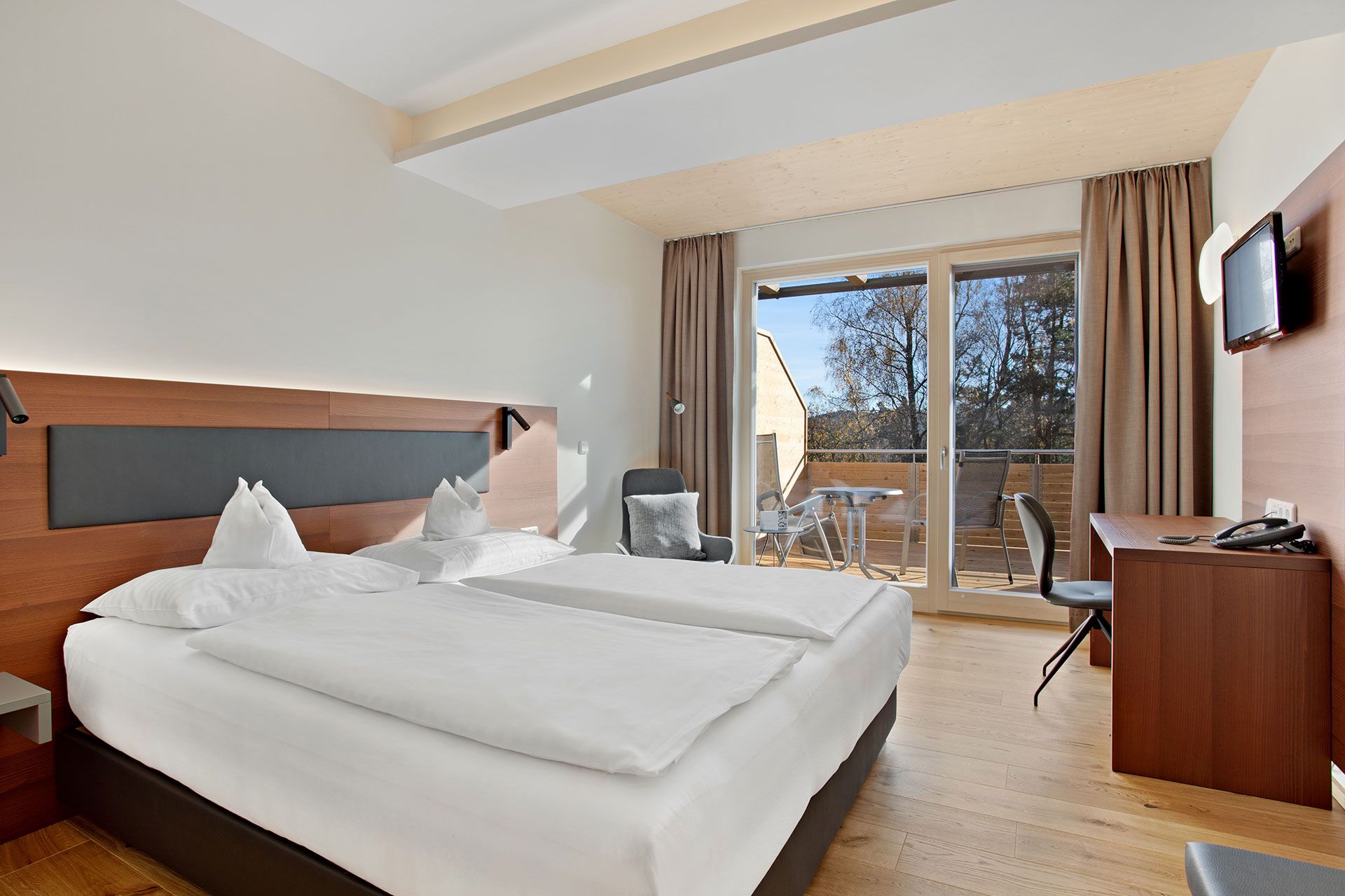 Modernes Hotelzimmer mit Doppelbett, Schreibtisch, Fernseher und Balkon mit Blick ins Freie.