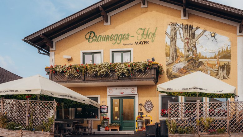 Ein traditionelles Gasthaus mit Balkon und Wandgemälde, umgeben von Sonnenschirmen und Pflanzen.