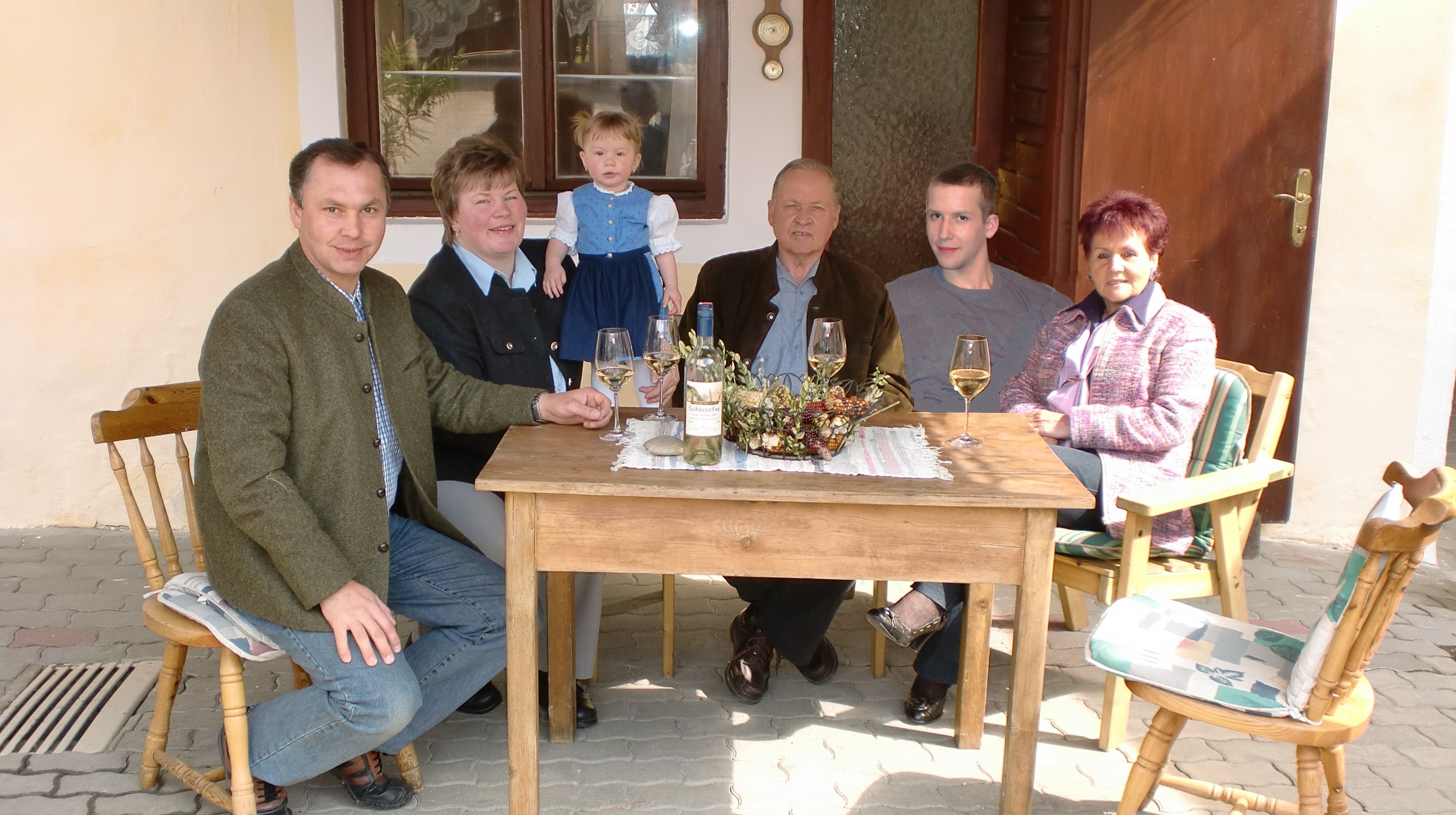 Eine Familie sitzt an einem Holztisch im Freien und trinkt Wein.